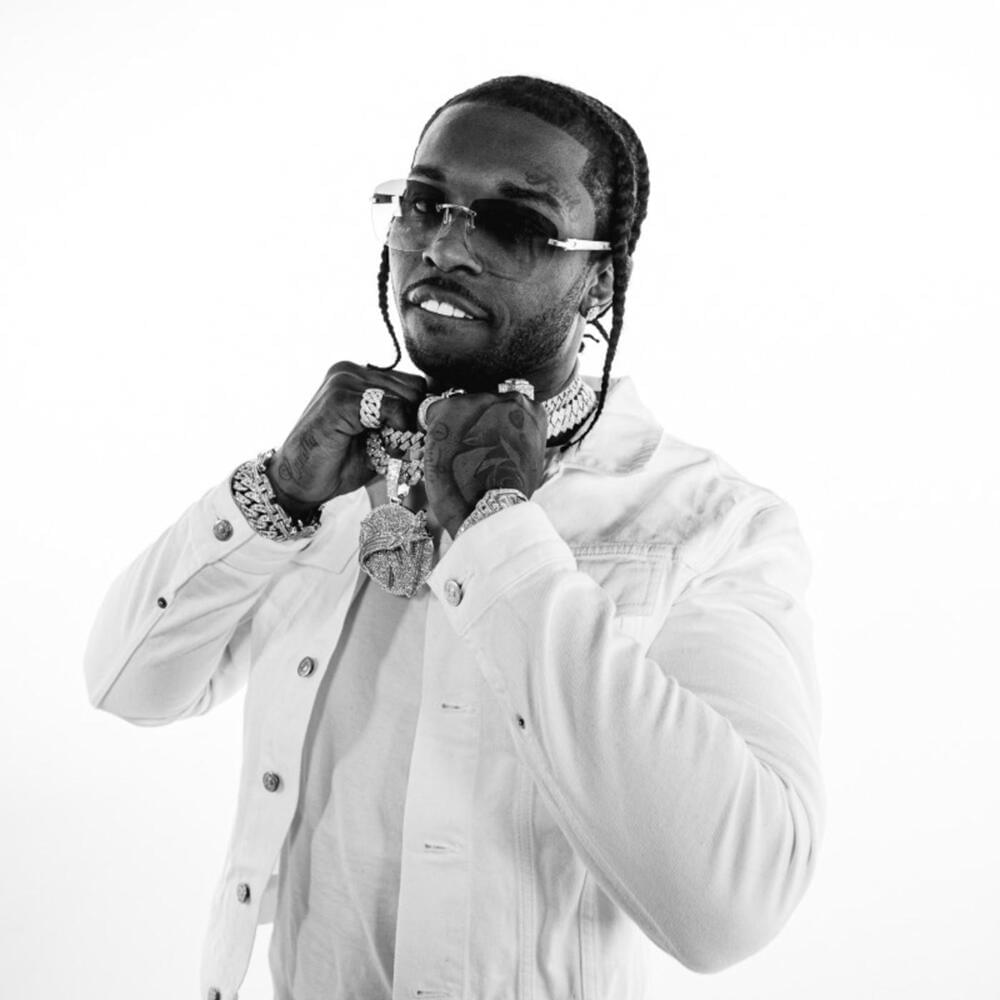 Pop Smoke wearing a white T-shirt and jacket, holding his chains with both hands while smiling.